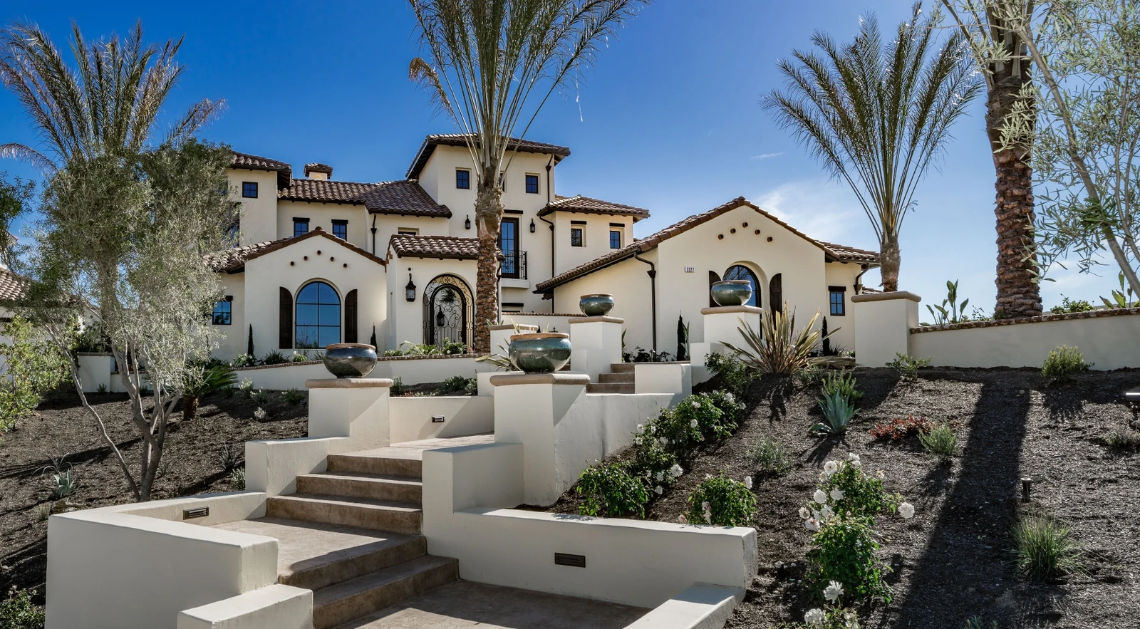 Custom-built Mediterranean-style residence with tile roof, arched entry, and terraced stone stairway surrounded by palms and drought-tolerant landscaping.